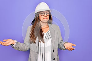 Professional woman engineer wearing industrial safety helmet over pruple background clueless and confused expression with arms and