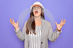 Professional woman engineer wearing industrial safety helmet over pruple background crazy and mad shouting and yelling with