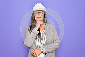 Professional woman engineer wearing industrial safety helmet over pruple background with hand on chin thinking about question,