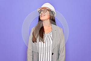 Professional woman engineer wearing industrial safety helmet over pruple background looking away to side with smile on face,