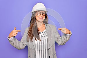 Professional woman engineer wearing industrial safety helmet over pruple background looking confident with smile on face, pointing
