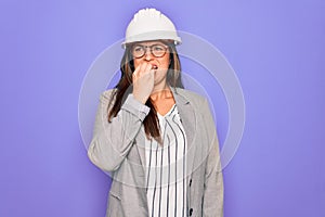 Professional woman engineer wearing industrial safety helmet over pruple background looking stressed and nervous with hands on