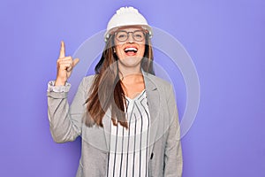 Professional woman engineer wearing industrial safety helmet over pruple background pointing finger up with successful idea