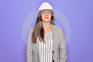 Professional woman engineer wearing industrial safety helmet over pruple background with serious expression on face