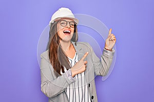 Professional woman engineer wearing industrial safety helmet over pruple background smiling and looking at the camera pointing