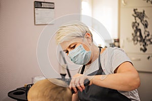 Professional woman hairdresser with face mask at work. Process of hair cutting at beauty salon. A boy gets a trendy haircut.