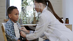 Professional woman pediatrician examining breathing system of little black boy patient with stethoscope and tickling him