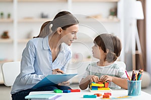 Professional woman psychologist exercising with little boy, checking readiness for school and taking notes at office