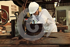 Professional young Asian worker in white uniform and safety equipment cutting a piece of wood on vertical band saw machine in carp