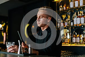 Professional young barman standing behind bar counter and holding stirring spoon and glass filled with ice cubes