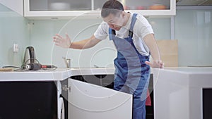 Professional young carpenter examining broken kitchen cabinet door in apartment. Brunette Caucasian service man in blue