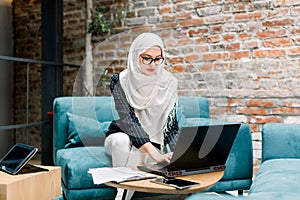 Professional young muslim business woman, wearing white hijab, using laptop computer, sitting on blue sofa in modern