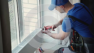 A professional young repairman in a special blue uniform with tools installs radiators and a thermostat in the apartment