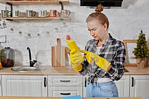 Professional young woman wearing rubber protective yellow gloves holding bottle cleaners in the kitchen.