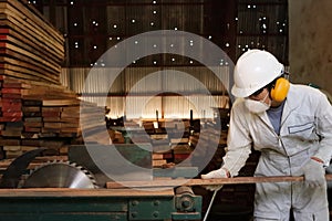 Professional young worker in white uniform and safety equipment cutting a piece of wood on table saw machine in carpentry factory.