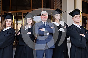 A professor and his students in graduation gowns stand with their arms crossed over their chests.