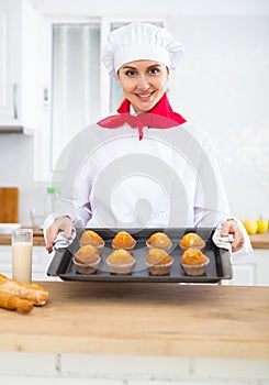 Proffesional woman cook in white uniform holding sheet pan with just baked cupcakes photo