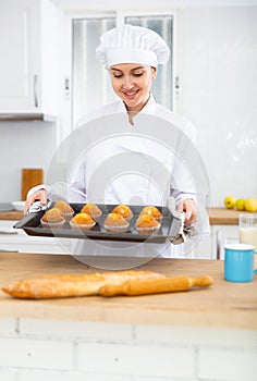Proffesional woman cook in white uniform holding sheet pan with just baked cupcakes photo