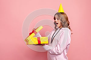 Profile of amazed happy teenage girl in hoodie opening gift box with opened mouth, satisfied with present