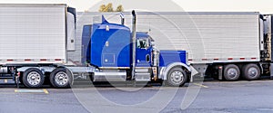 Profile of big rig blue semi truck with reefer semi trailer standing in row with another semi trucks on truck stop