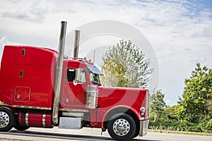 Profile of bright classic red big rig semi truck tractor with vertical chrome pipes running on the green highway