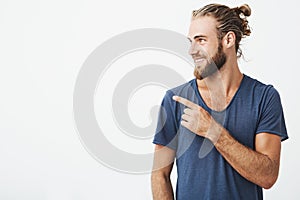 Profile of cheerful handsome man with fashionable hairstyle and beard smiling brightfully and pointing at free space for