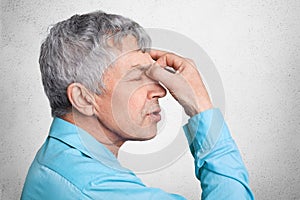 Profile of exhausted grey haired male in formal shirt, keepd hands on nose, poses against white concrete background, tries to conc