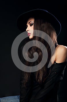 Profile of expressive female model posing in black shirt and elegant hat with red bright lipstick on dark shadow background.