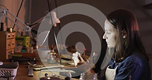 Profile of focused caucasian female jeweller sitting at desk, making jewelry in workshop