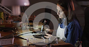 Profile of focused caucasian female jeweller sitting at desk, making jewelry in workshop