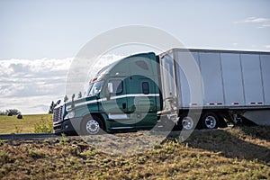 Profile of green big rig semi truck with dry van semi trailer running on the road through the fields
