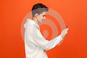 Profile of happy smiling teenager in casual hoodie holding mobile phone in hands chatting via video call, navigation application