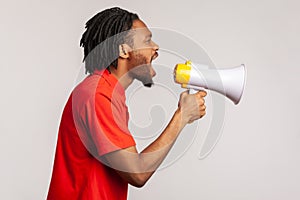 Profile of man with dreadlocks and beard wearing red casual style T-shirt, loudly screaming at megaphone, making protest, wants to