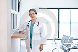 Profile photo of attractive smiling female doctor wearing white medical gown with stethoscope, standing in hospital corridor.