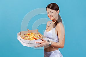 Profile portrait attractive slim woman in white sports top holding cardboard box with fatty hot pizza, looking at camera with