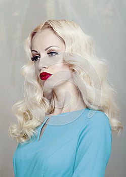 Profile portrait of a beautiful young blonde woman with curly hair and makeup, red lips, dressed in a blue dress.