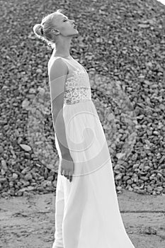 Profile portrait of blonde woman with hair tight up in white bride dress, has got closed eyes, over piles of stones.