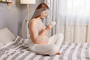 Profile portrait of cute beautiful pregnant brown haired caucasian woman sitting in bed holding cup of hot beverage enjoying tasty