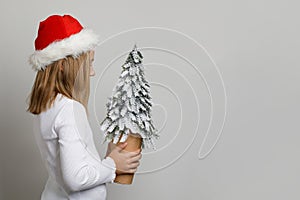 Profile portrait of cute child girl in red Santa hat holding snowy green Christmas tree on white studio wall banner background