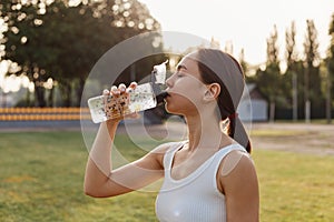 Profile portrait of fe male with dark hair and ponytail wearing white top posing outdoors and drinking water from bottle, being