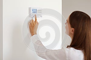 Profile portrait of female with dark hair wearing white casual style shirt adjusting temperature on thermostat, pressing buttons,