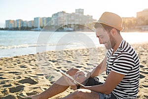 Happy man in hat sitting on beach using tablet