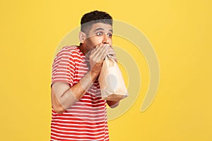 Profile portrait scared man in striped red t-shirt exhaling and inhaling into package, using paper bag to improve well-being,