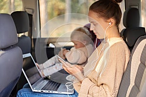 Profile portrait of serious woman with earphones and mobile phone working on laptop while sitting with her toddler kid in safety