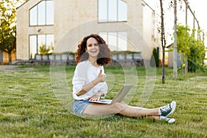 Profile portrait of a smiling woman with curly hair with laptop seated on grass in the her yard, points his thumb up