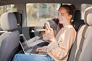 Profile portrait of woman wearing beinge jumper sitting with her baby daughter in safety chair on backseat of car, holding mobile