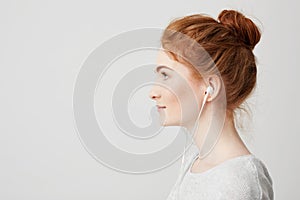 Profile portrait of young beautiful tender redhead girl with bun in headphones smiling over white background.