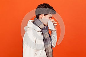 Profile portrait of young man in casual hoodie with checkered scarf on neck blowing his nose into napkin, seasonal allergy
