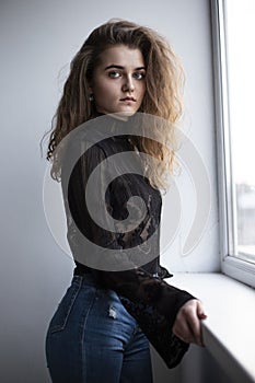 Profile portrait of a young woman with curly brown hair, dressed in a black suitcase and jeans, posing near window.