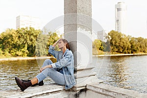 Profile of a serious single pensive teen girl, dressed in denim, seated outside near river, looking away.